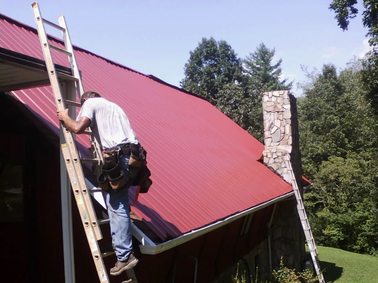 Roofer on a ladder beside a red metal roof and stone chimney — Soffit & Fascia Repair by Roofing Brothers in Willards